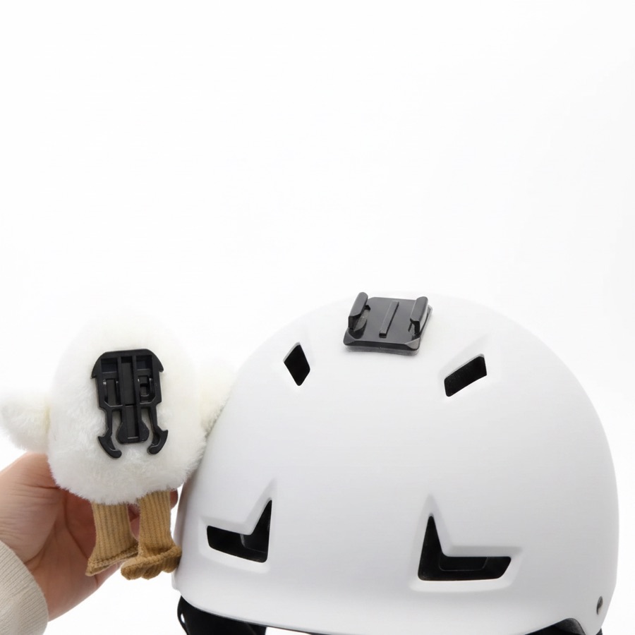 A plush topper turned over to show its built-in buckle clip, held next to a helmet with the peel-and-stick mount already attached to its crown